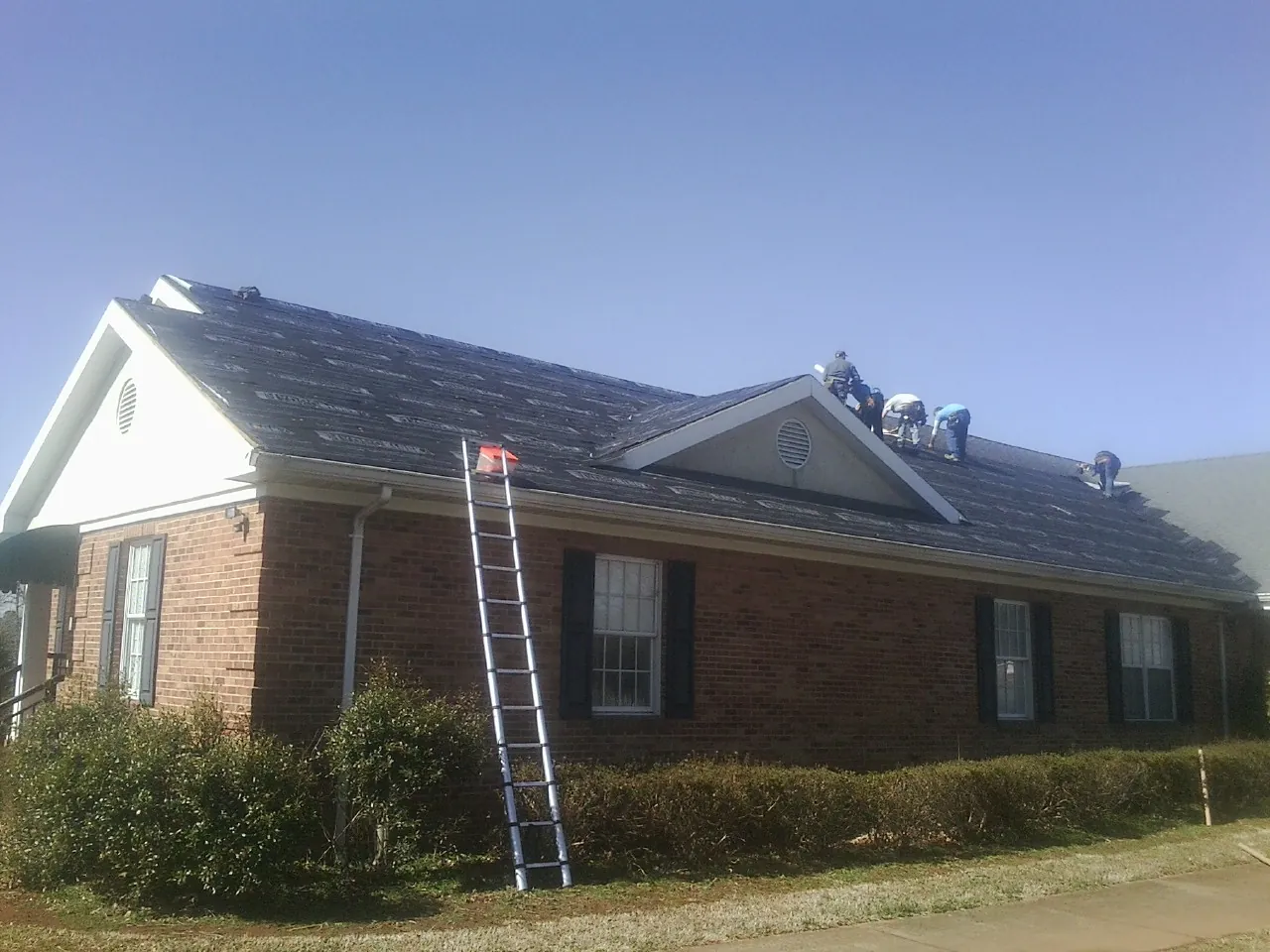 Crew tearing shingles off a brick ranch roof with a ladder set — Metal Roof Repair work by our crew in Erhard