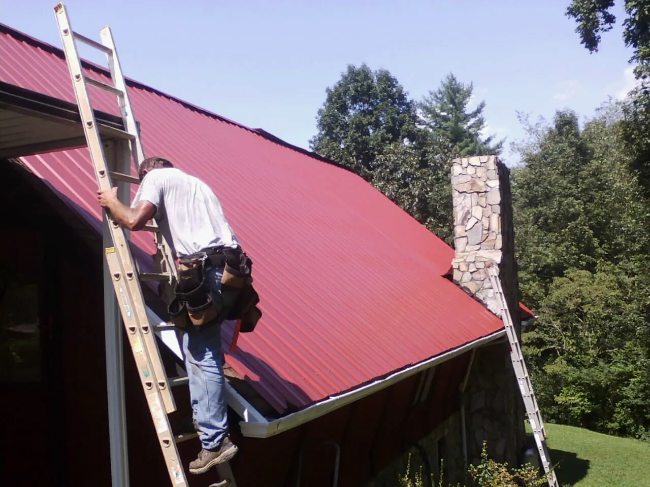 Roofer on a ladder beside a red metal roof and stone chimney — Soffit & Fascia Repair by Roofing Brothers in Erhard